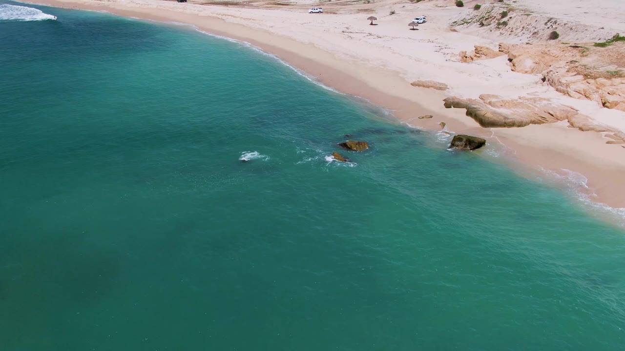 Aerial view at El Tule Los Cabos showing turquoise ocean water beside a pale sandy beach, with scattered shoreline rocks and no visible people.
