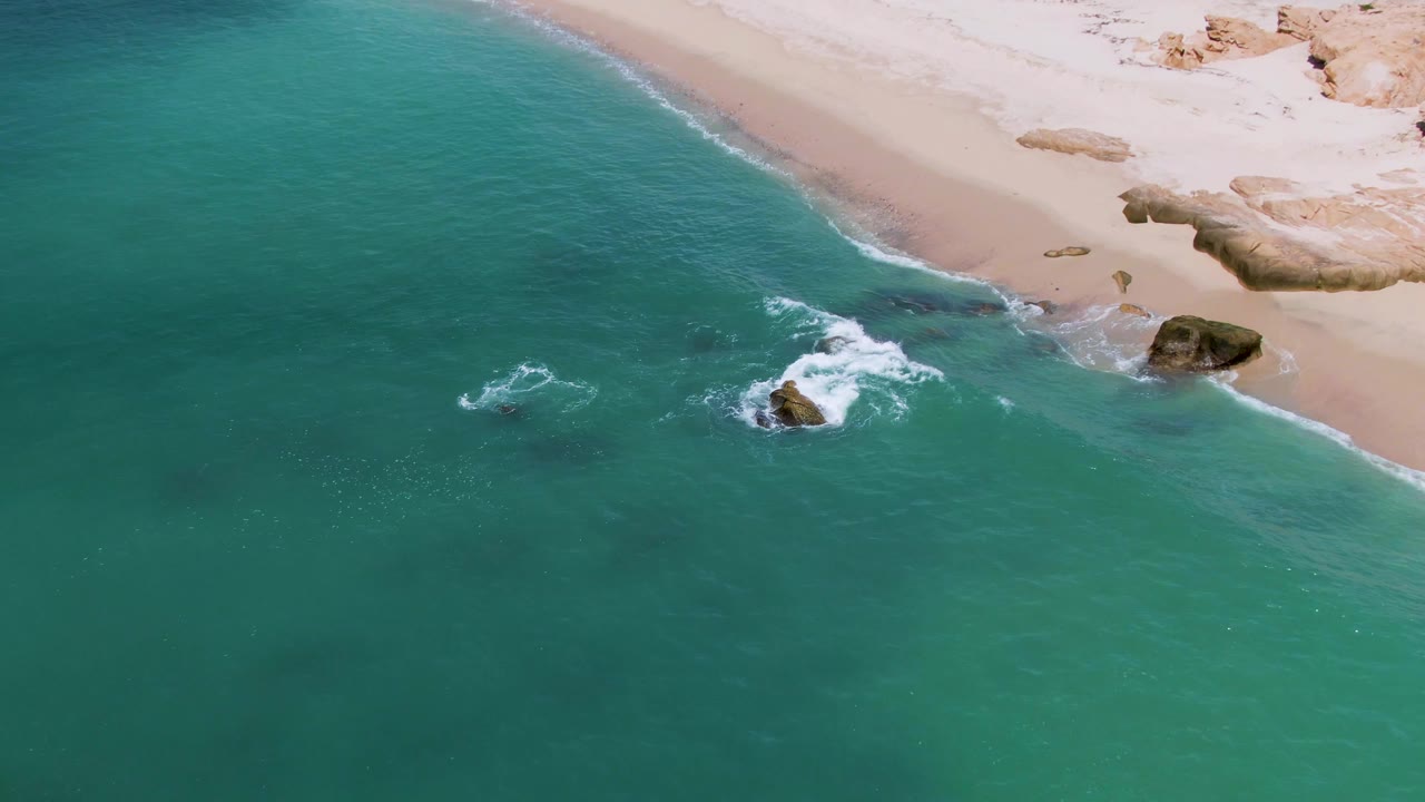 Aerial view at El Tule Los Cabos where white surf curls around nearshore rocks below a pale sand beach, with no visible people.