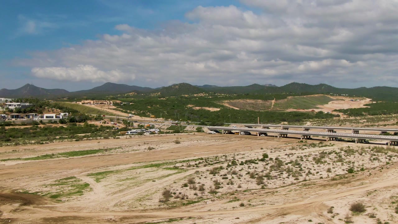 Aerial view in El Tule Los Cabos showing a dry sandy riverbed in front, a bridge with small vehicles in the distance, green hills, mountains, and no visible people.
