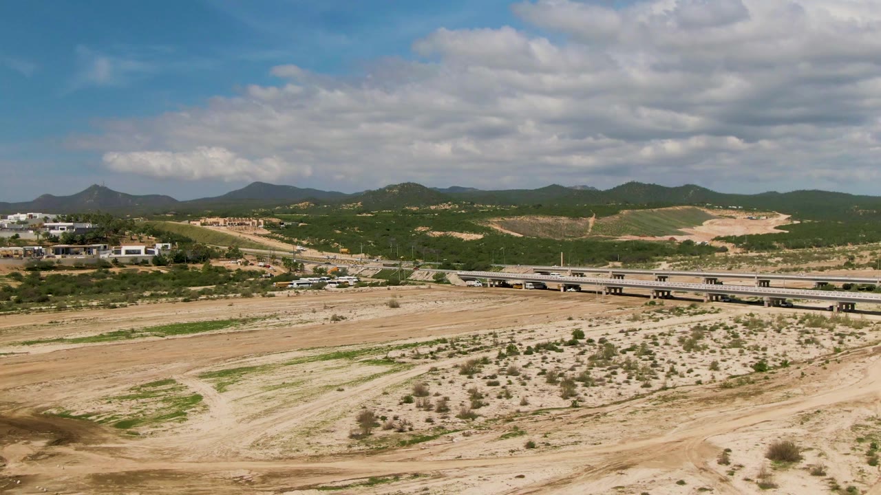 A wide aerial frame in El Tule Los Cabos shows the sandy riverbed stretching across the foreground, the bridge centered farther back, hillside buildings at left, and no visible people.
