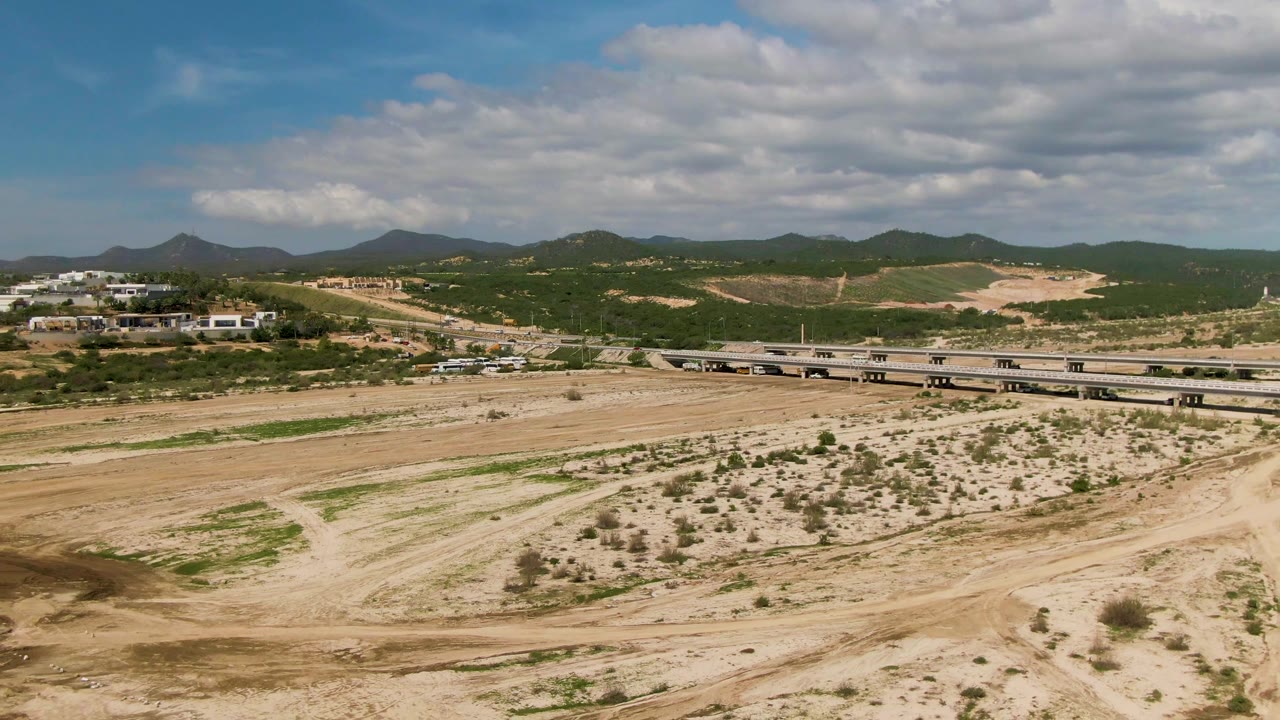 In El Tule Los Cabos, the frame places the dry riverbed across the lower half, a bridge with several vehicles through the midground, white buildings at left, and no visible people.