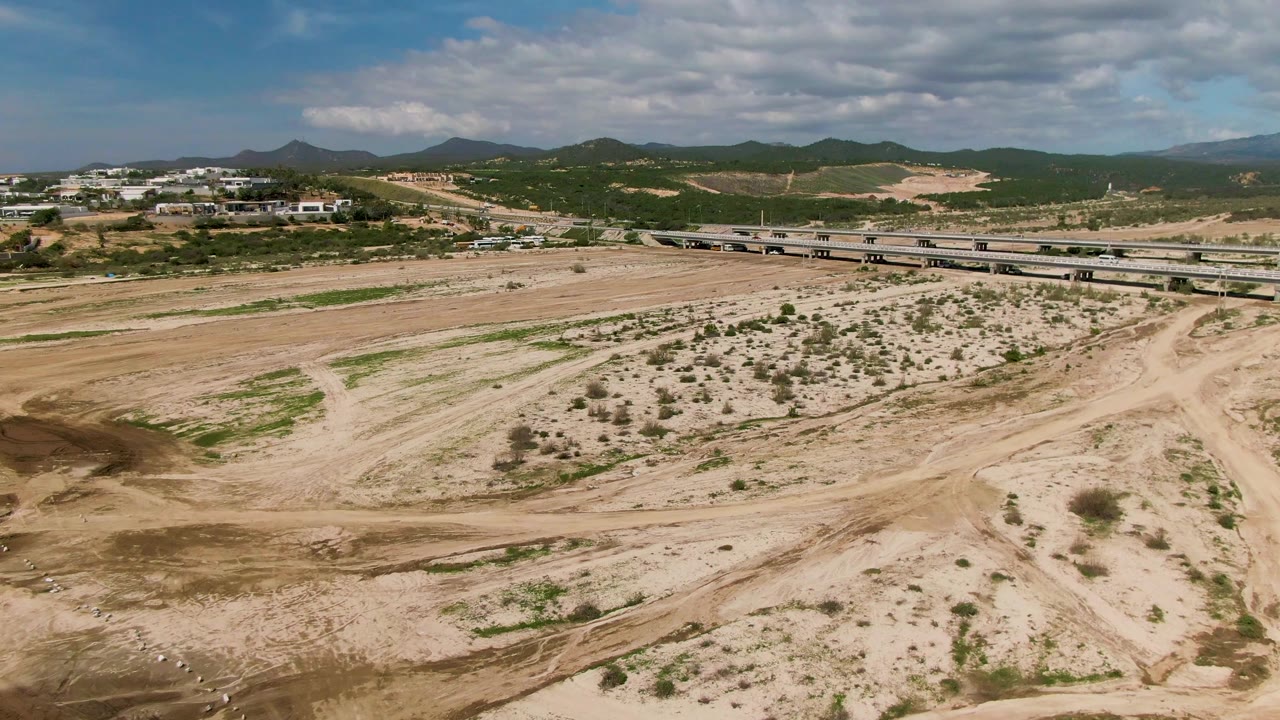 Aerial view in El Tule, Los Cabos, with a bridge spanning dry sandy ground, sparse shrubs, distant buildings, green hills, cloud cover, and no visible people.