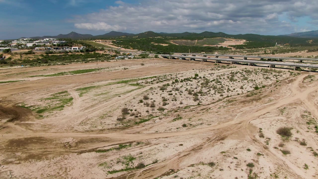 Aerial view in El Tule, Los Cabos, with the bridge extending across the frame, sandy ground and scattered brush below, low hills and buildings in the background, and no visible people.