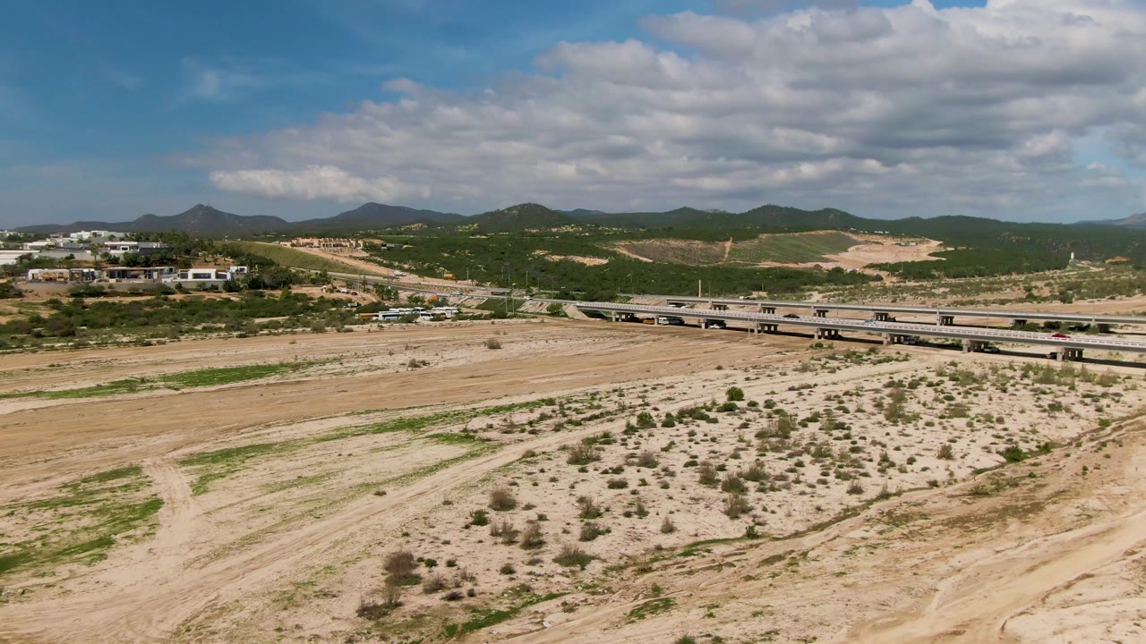 Aerial view in El Tule Los Cabos, Baja California Sur, showing vehicles on a low bridge over sandy ground with shrubs, distant hills, and no visible people.