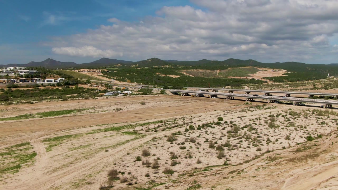 The bridge runs across the midground in El Tule Los Cabos with small vehicles visible, sandy foreground tracks, green hills, and no visible people.