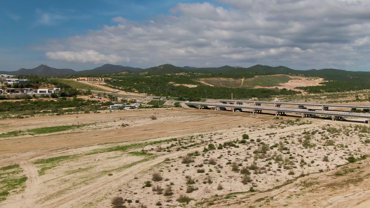 A wide aerial frame shows vehicles crossing the bridge near El Tule Los Cabos, with scrubby sand flats in front, layered green hills behind, and no visible people.
