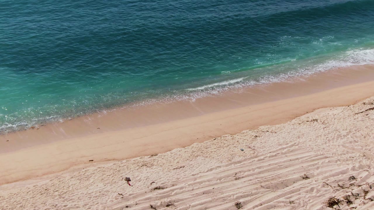 Aerial frame from El Tule Los Cabos showing tan sand below, a thin white wave line at the shore, turquoise shallows, darker blue water beyond, and a small person on the beach.