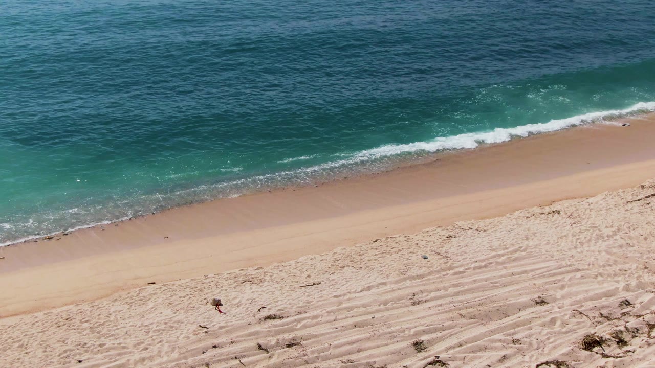 Aerial view over El Tule Los Cabos with pale sand, a narrow foamy break, clear turquoise nearshore water, darker ocean beyond, and small people visible near the beach and waterline.