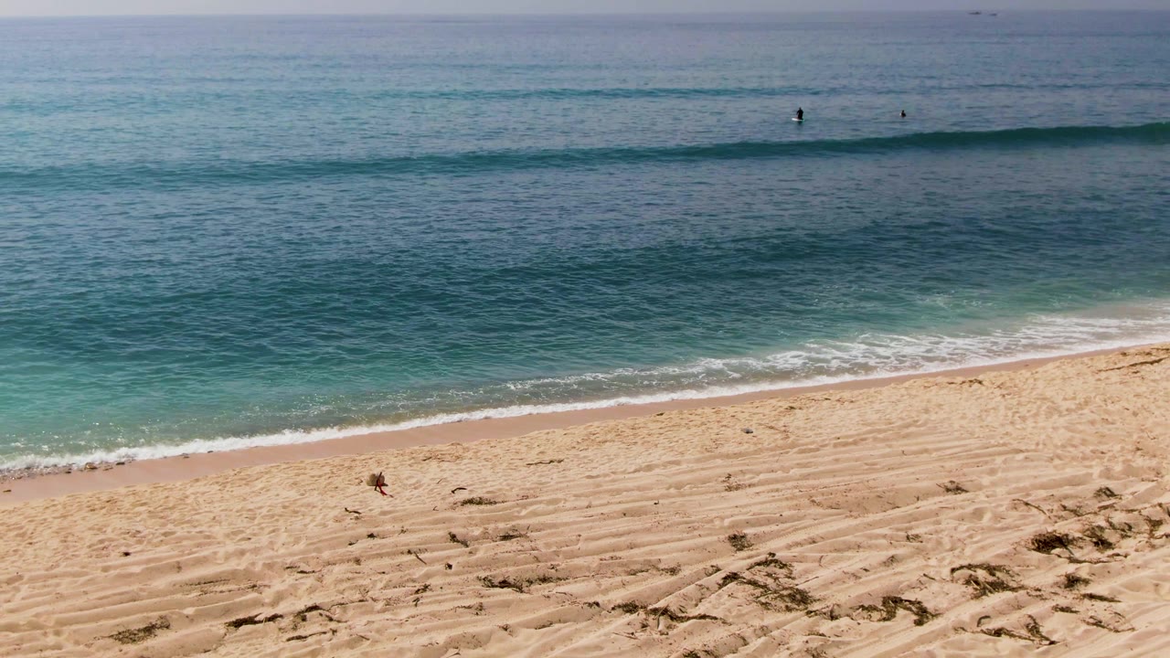 Wide view of El Tule Los Cabos beach with rippled sand in front, turquoise water beyond, and a few distant swimmers near a low wave line.