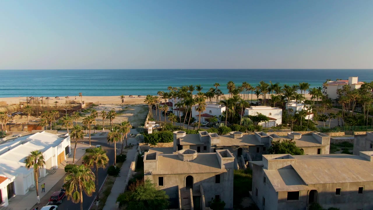 Opening frame of San Jose Del Cabo coastal residences with flat rooftops in the foreground, palms in the midground, beach and ocean beyond, and no visible people.