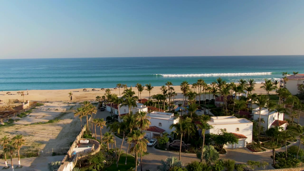 Aerial view in San Jose Del Cabo with more open sand visible at left, palms and white buildings below, and a low surf line across the blue water with no visible people.