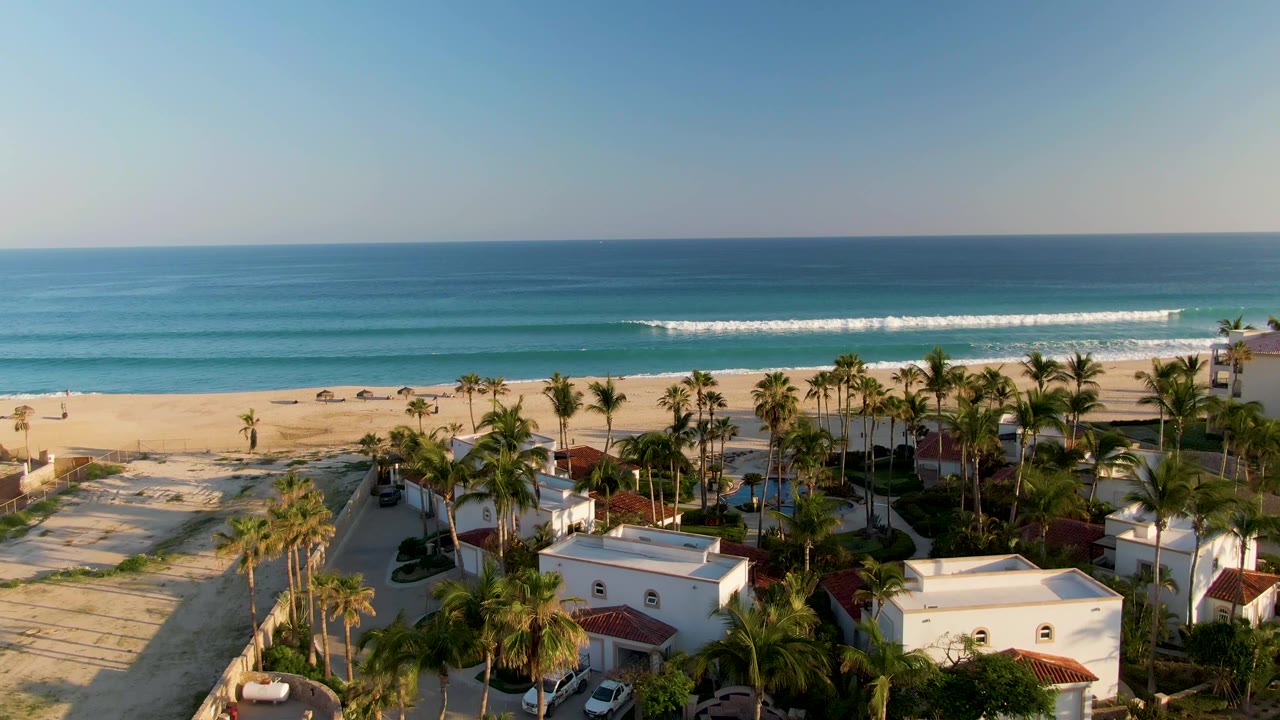 San Jose Del Cabo aerial frame with palm crowns and white residences across the lower edge, a sandy beachfront in the middle, and turquoise-blue water under clear sky with no visible people.