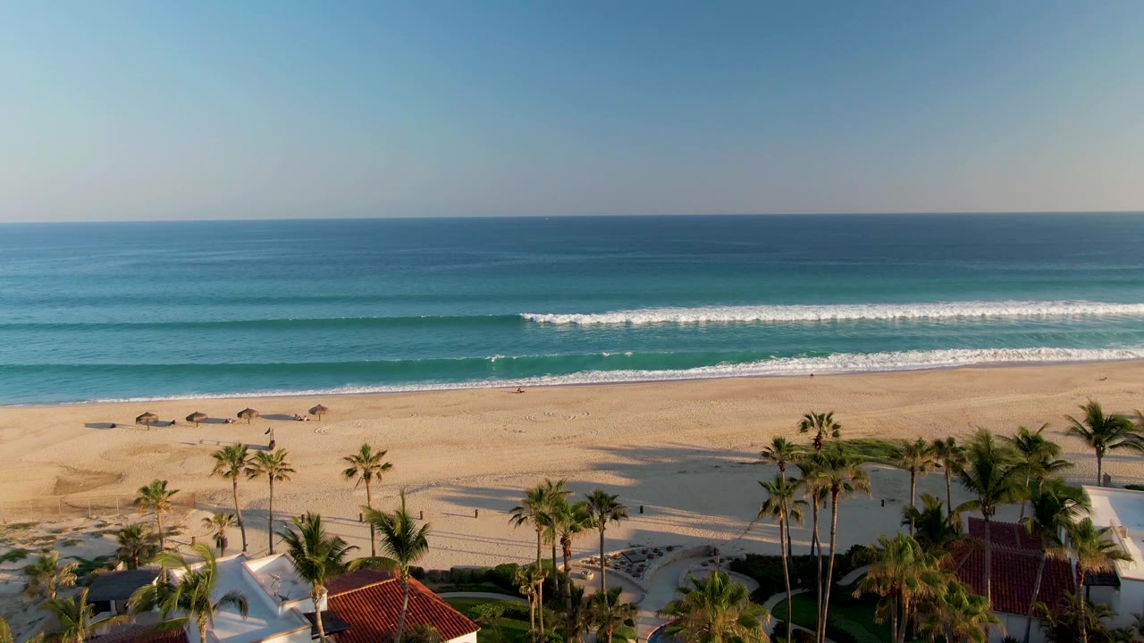 Opening frame of San Jose del Cabo beach with palms and small rooftops at the lower edge, tan sand, white surf lines, blue ocean, clear sky, and no visible people.