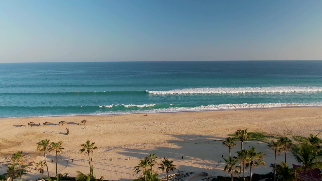 San Jose del Cabo beach frame with a lower strip of palms and roof edge, broad sand, small shade structures, rolling white waves, blue water, clear sky, and no visible people.