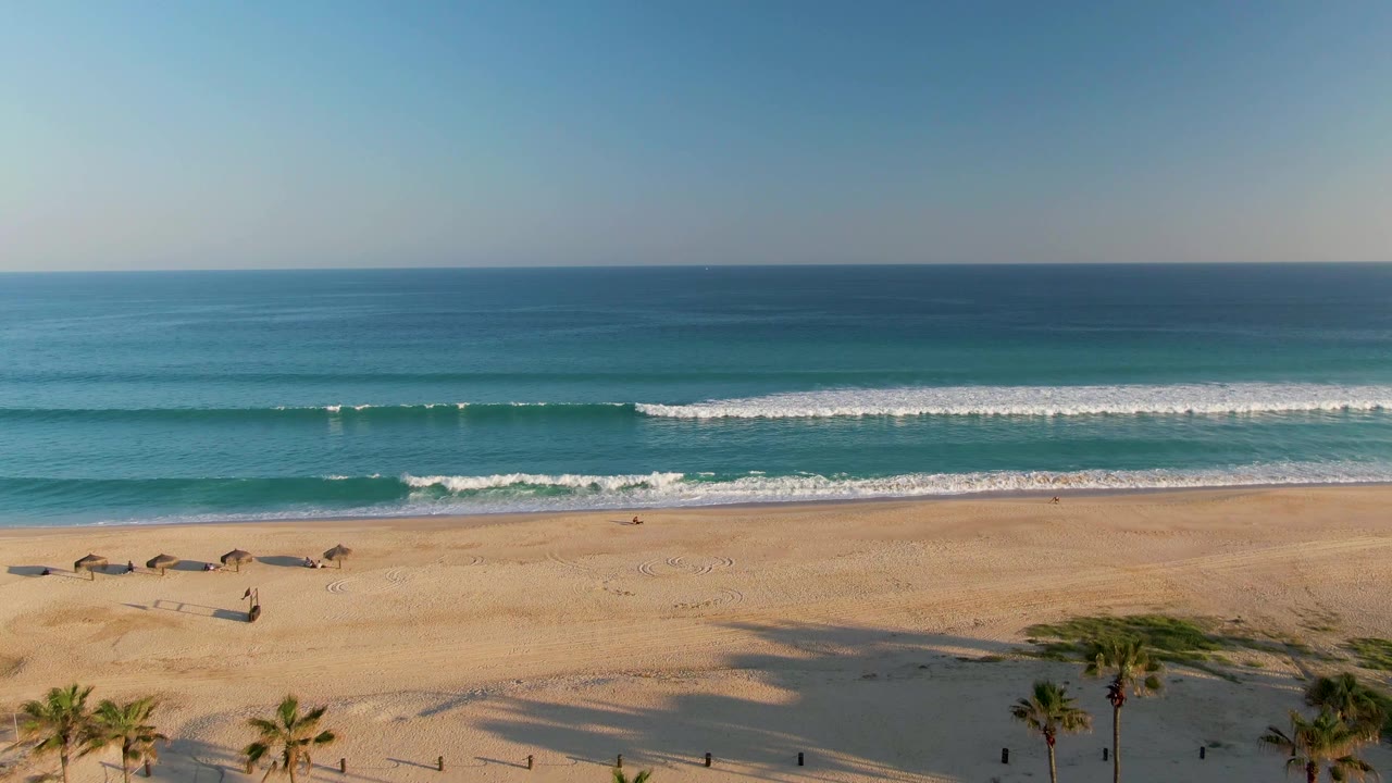 San Jose del Cabo shoreline with blue ocean, a long white wave band, open tan sand, clustered palms near the lower edge, clear sky, and no visible people.