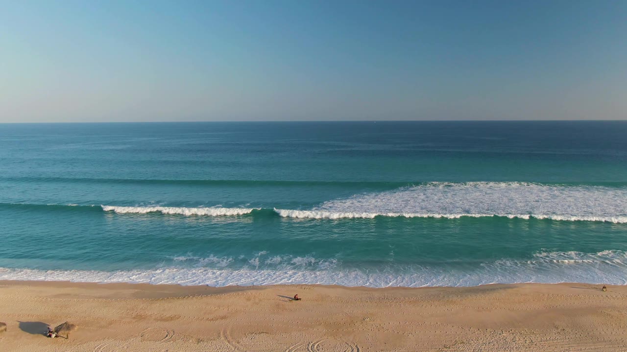 San Jose del Cabo shoreline frame with a long white surf line breaking across blue-green water and a narrow strip of tan beach below.