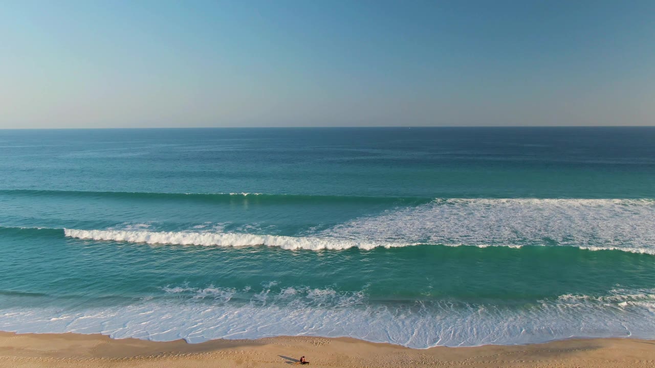 San Jose del Cabo ocean frame with a broad white surf band rolling toward the lower beach edge and a clear horizon above the blue water.