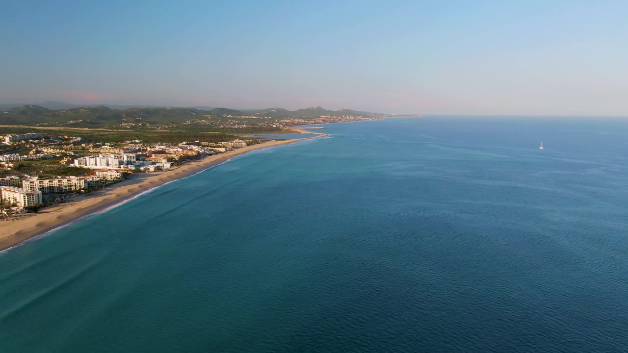 San Jose del Cabo coastline seen from above with the beach and buildings on the left, open blue water to the right, faint wave lines near shore, and no visible people.