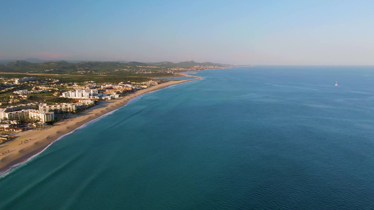 San Jose Del Cabo aerial frame showing the coast receding toward the horizon, low beachfront buildings beside the sand, blue ocean across the right side, and no visible people.