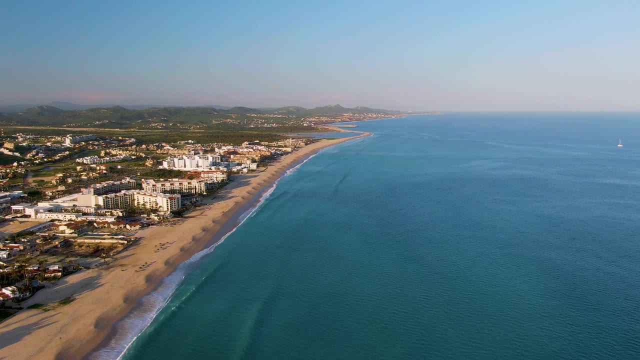 Aerial view of sandy beach, turquoise ocean, shoreline buildings, and a distant sailboat.