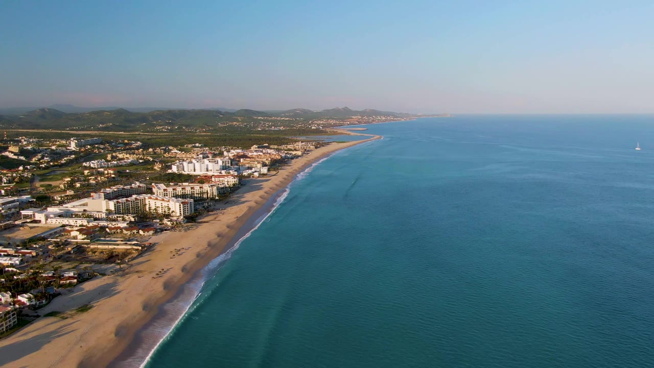 Aerial frame from San Jose Del Cabo with the shoreline running diagonally through the image, beach and buildings at left, open blue water at right, distant mountains, and no visible people.
