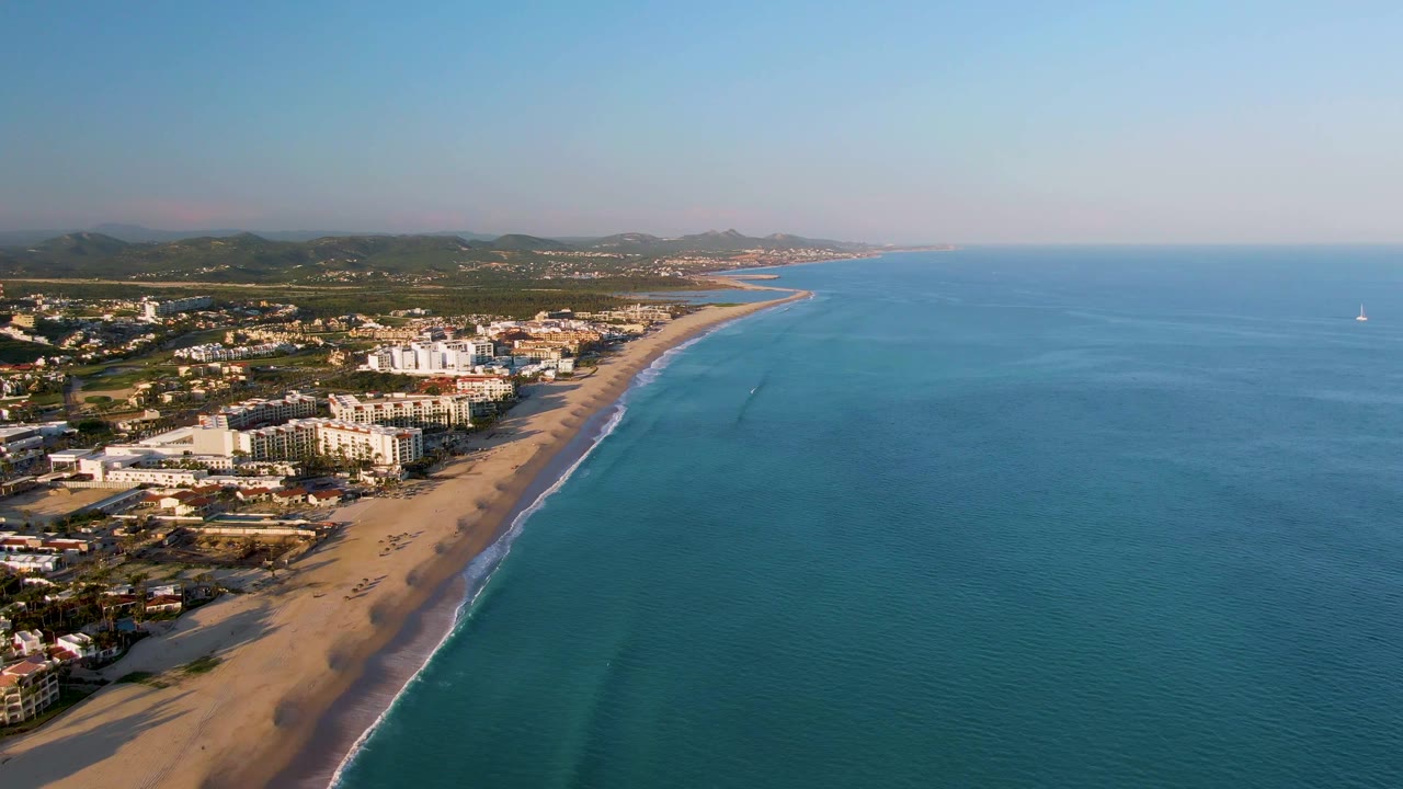 Aerial frame from San Jose Del Cabo showing more open ocean to the right, the sandy shoreline and buildings along the left edge, a hazy mountain horizon, and no visible people.