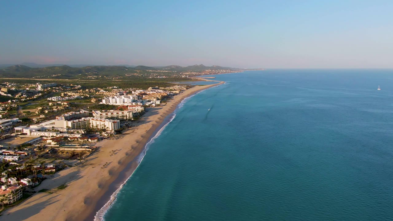 Aerial frame from San Jose Del Cabo with pale sand in the lower left, turquoise water filling the right side, shoreline buildings behind the beach, inland hills, and no visible people.