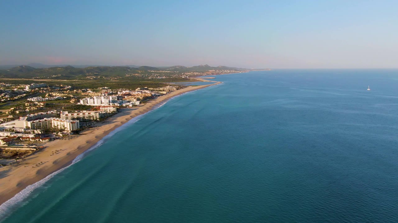 Opening aerial view of San Jose Del Cabo coastline with sandy beach and white beachfront buildings on the left, blue ocean on the right, low hills in the distance, and no visible people.