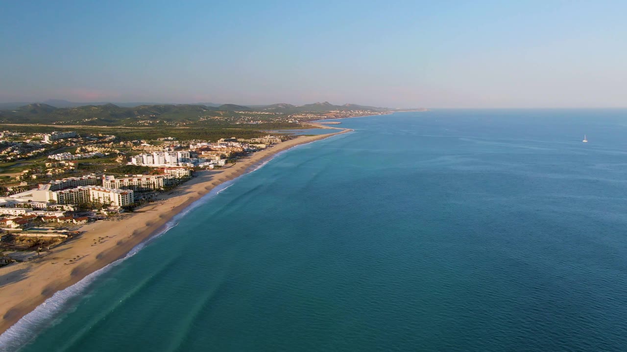Aerial frame over San Jose Del Cabo with the surf line running diagonally past beachfront buildings, open blue water filling the right side, a small sailboat offshore, and no visible people.