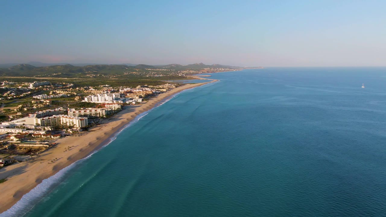 San Jose Del Cabo aerial still showing beachside buildings clustered near the shore, gentle wave bands along the sand, clear blue sea, distant hills, and no visible people.