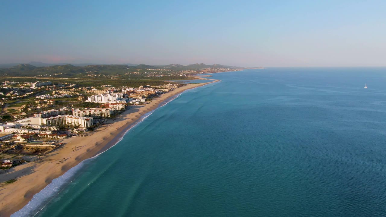Aerial view of sandy coastline with buildings, blue water, breaking waves, and hills.