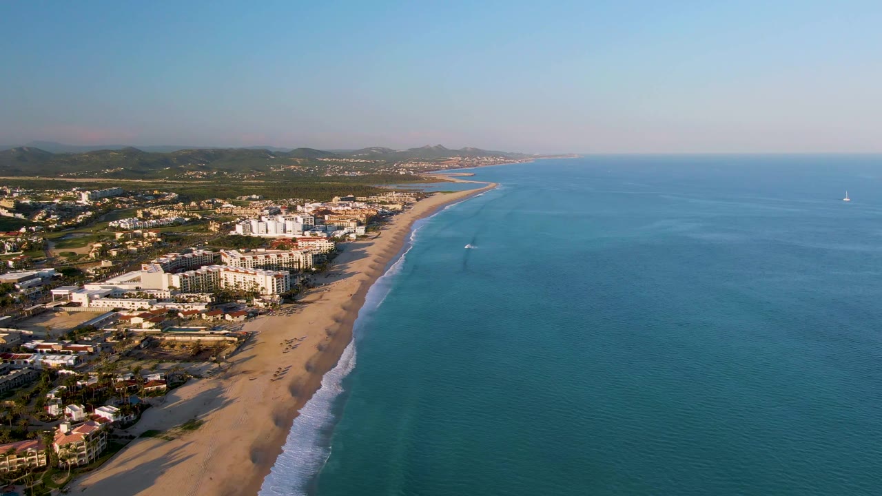 Aerial view over San Jose Del Cabo showing a sandy beach curving beside blue ocean, coastal buildings inland, white surf on the shore, and no visible people.