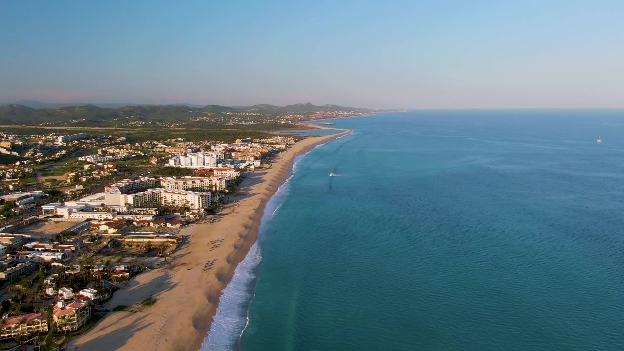 High aerial frame from San Jose Del Cabo with the sandy shoreline closer to center, white foam tracing the beach, a small watercraft offshore, and no visible people.