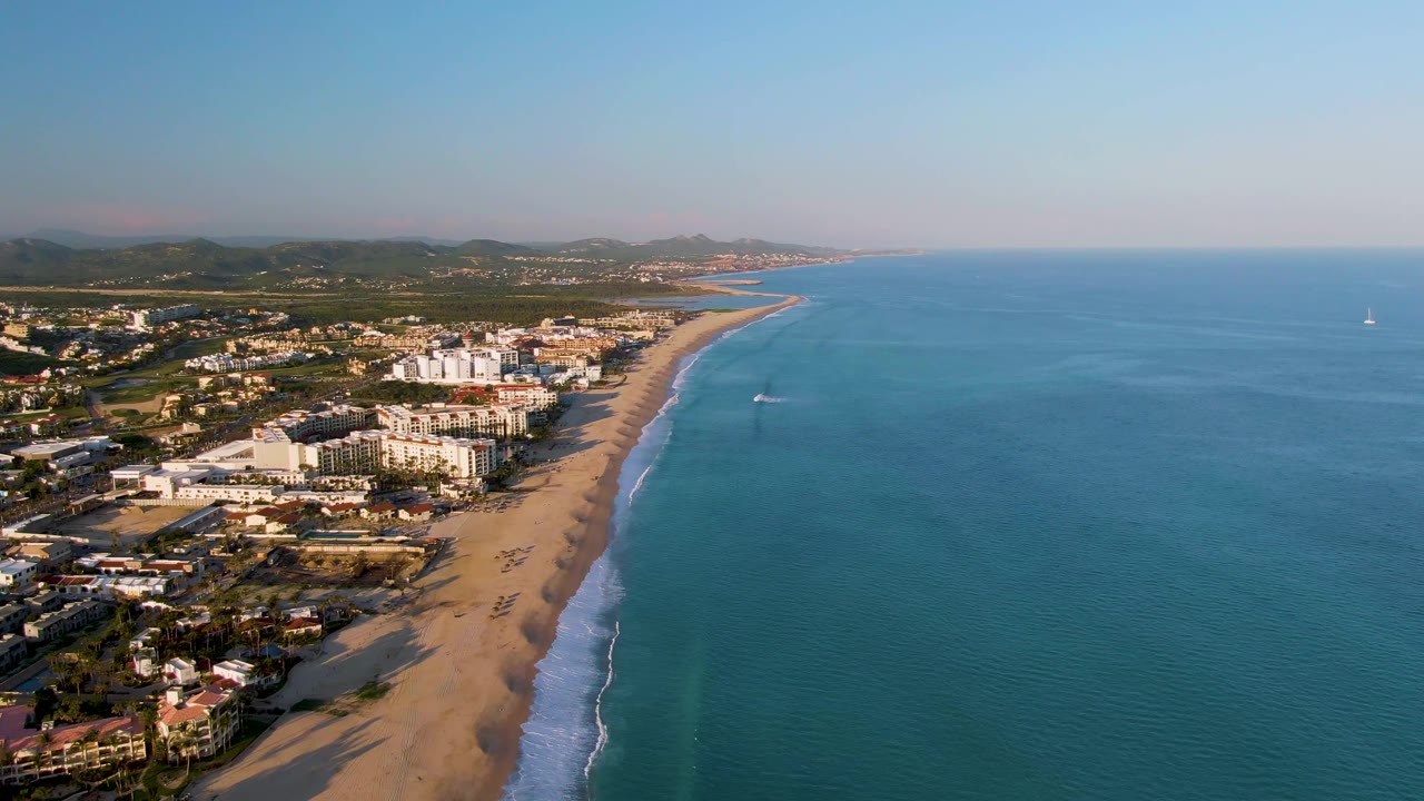 San Jose Del Cabo aerial frame showing beach buildings on the left, a long sandy shore below, open blue water on the right, a distant sailboat, and no visible people.