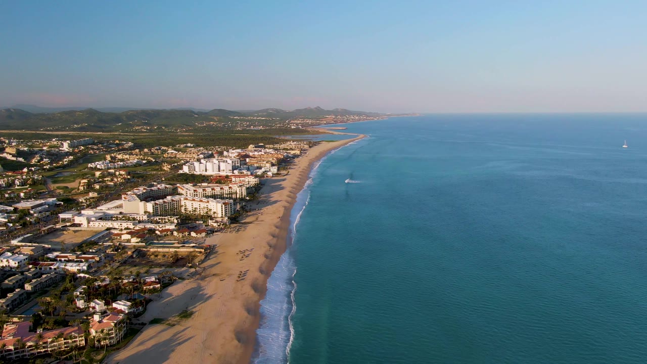 Aerial shoreline with beach homes or buildings, surf foam, blue water, and green inland terrain.
