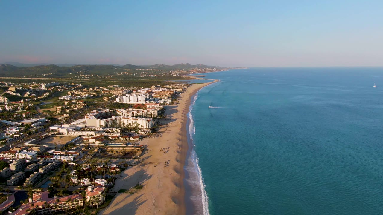 Aerial view over San Jose del Cabo showing a sandy beach beside coastal buildings, blue ocean to the right, a faint boat wake near shore, distant hills, and no visible people.