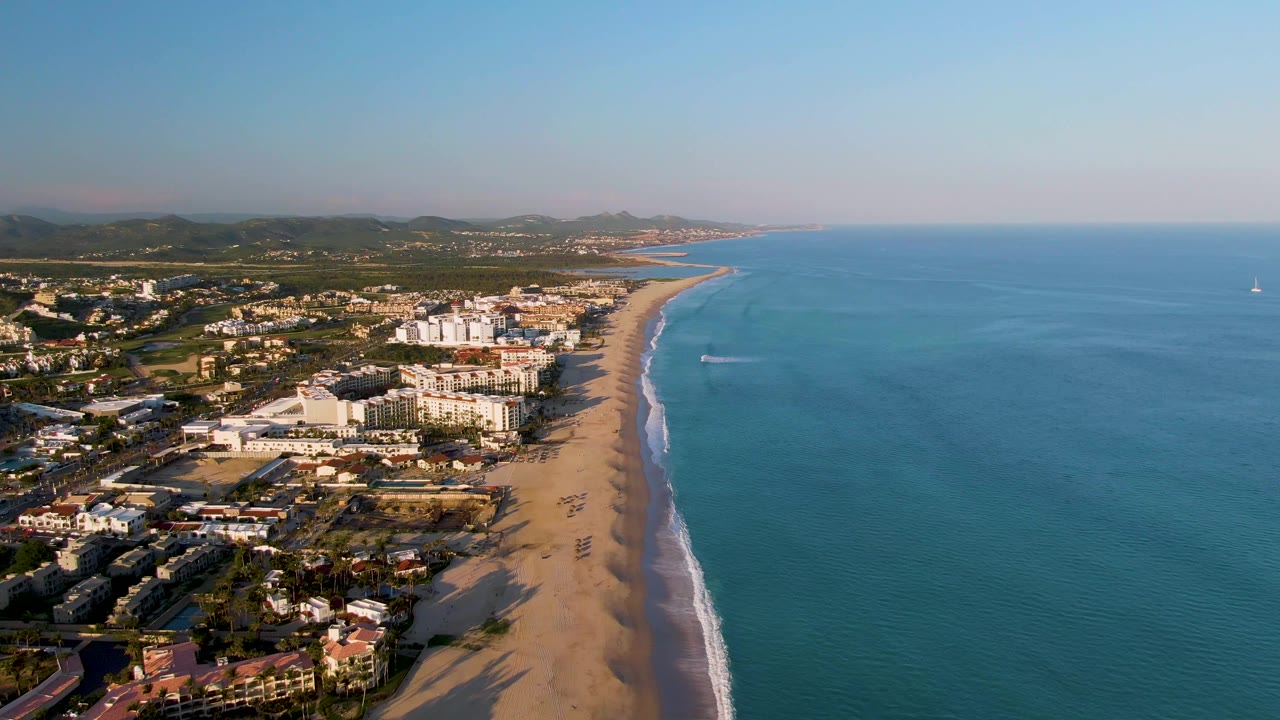 Drone view of San Jose del Cabo with the coastal town on the left, long sand beach below, blue water on the right, the wake line slightly farther along the shore, and no visible people.