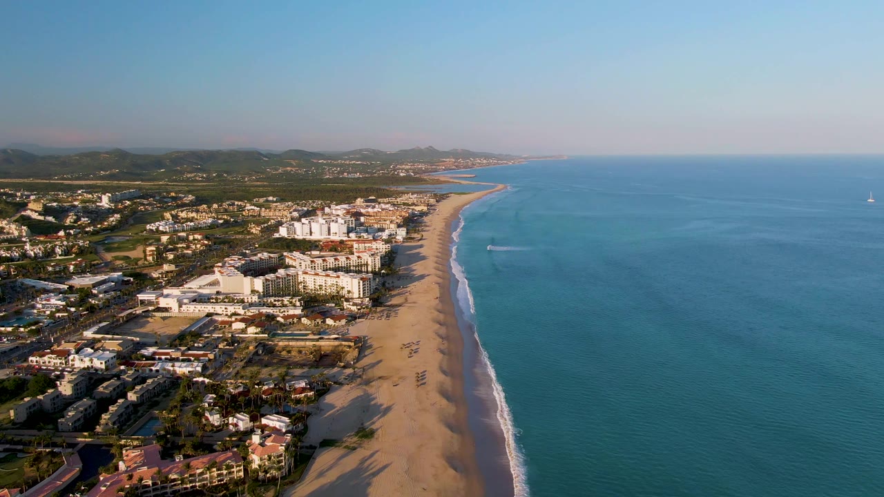 Elevated San Jose del Cabo coastline view showing beachside buildings, a broad band of sand, calm blue ocean, a small wake near the surf line, hazy hills inland, and no visible people.