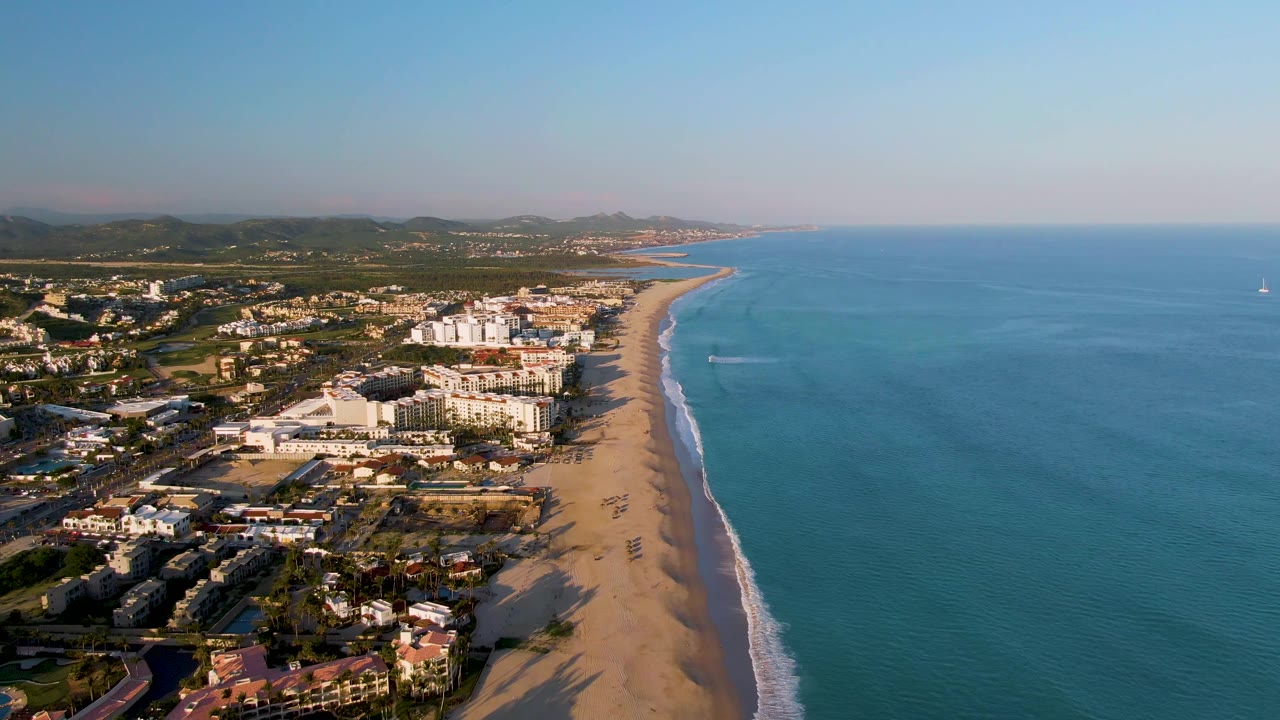 Aerial beach shoreline with town blocks, blue sea, distant hills, and fading boat wake.