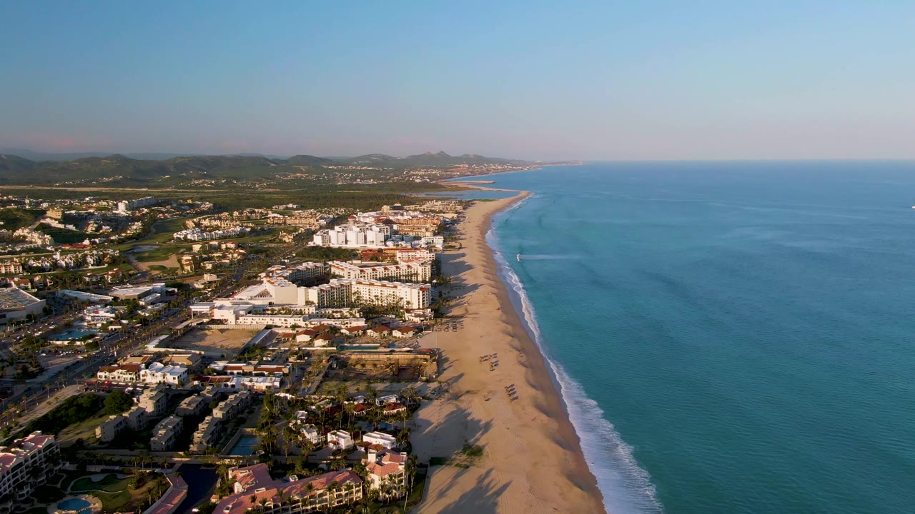 Aerial view of San Jose Del Cabo coastline at the start of the clip, with sandy beach running beside blue ocean, coastal buildings inland, distant mountains, and no visible people.