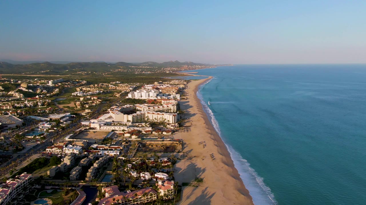 Wide aerial frame over San Jose Del Cabo with the beach curving into the distance, ocean on the right, clustered coastal buildings on the left, and no visible people.