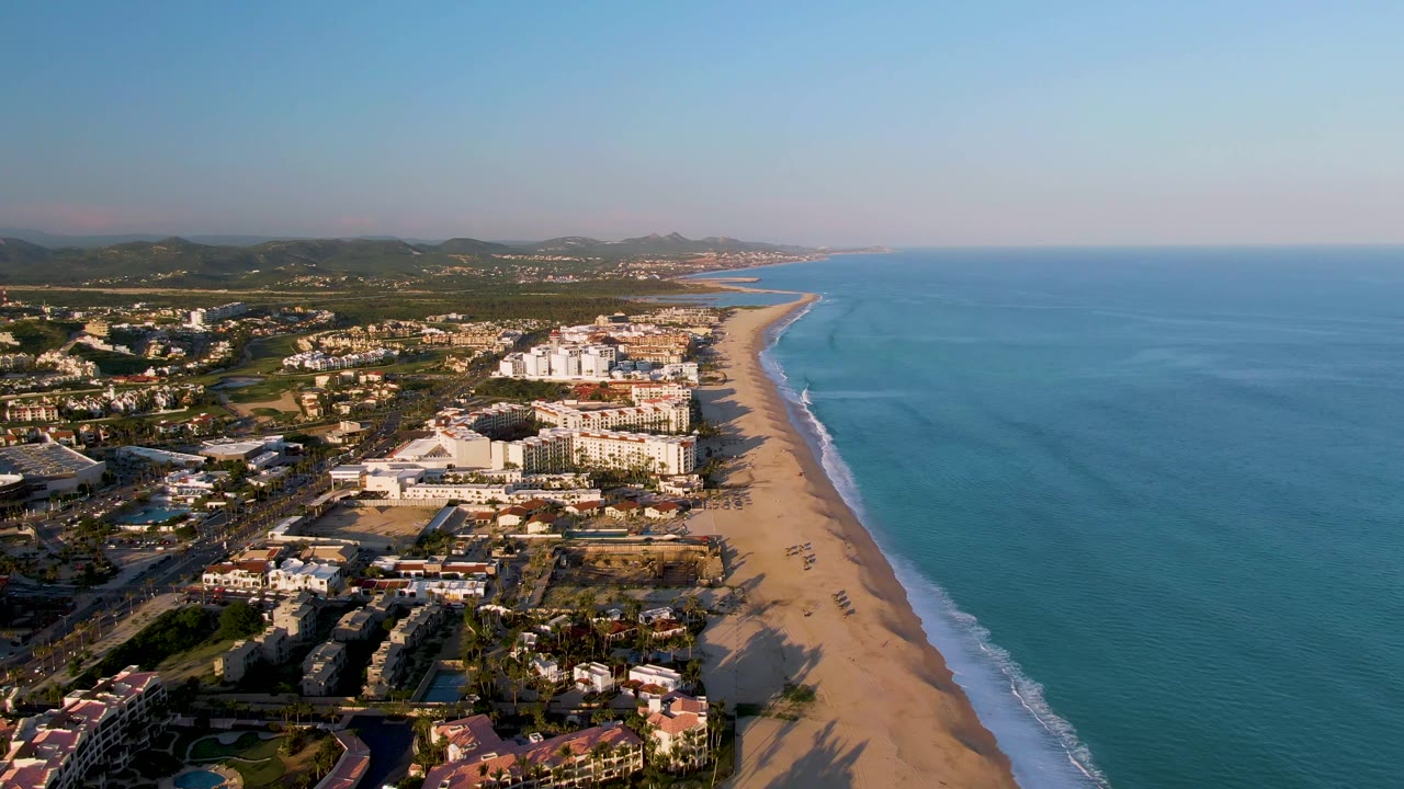 High aerial view in San Jose Del Cabo showing hotels or coastal buildings near the sand, a narrow surf line, open blue water, inland greenery, mountains, and no visible people.