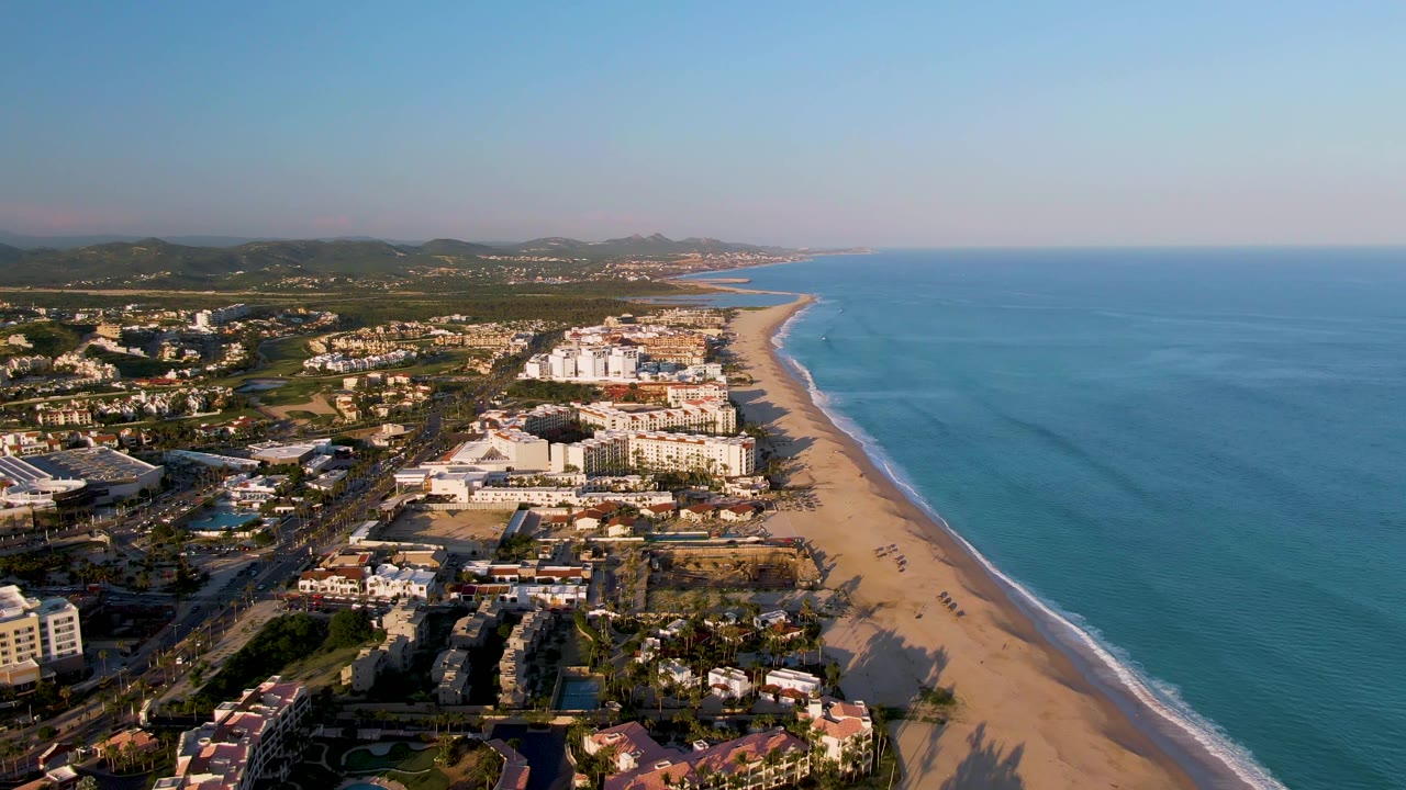 Aerial frame over San Jose del Cabo, Baja California Sur, with a diagonal sandy beach, blue ocean to the right, resort buildings and roads inland, low hills in the distance, and no visible people.