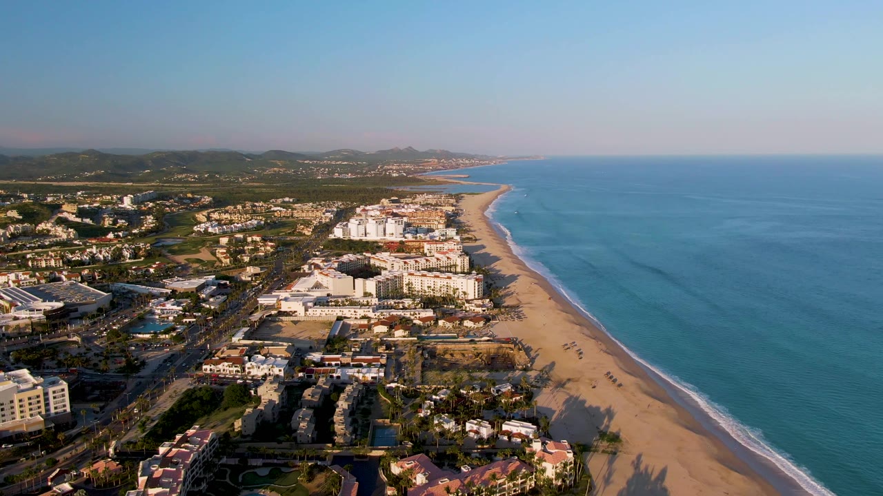 Second aerial frame of San Jose del Cabo coastline showing the resort district centered in frame, the sandy shoreline angling toward the background, blue water on the right, and no visible people.