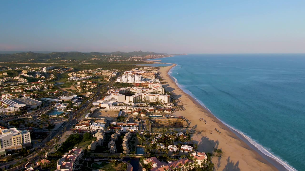 Aerial frame over San Jose del Cabo with the coastline stretching into the distance, hotel and resort structures clustered near the beach, open blue ocean filling the right side, and no visible people.