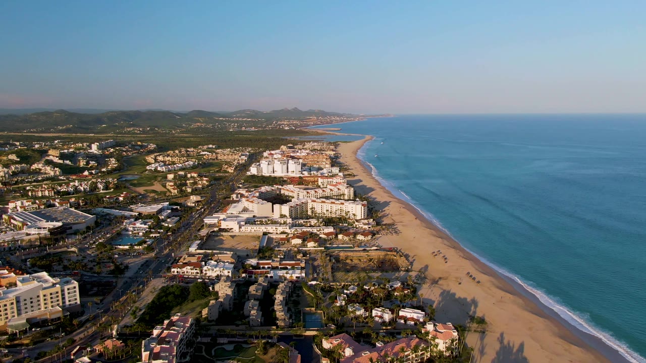 Wide aerial of beach resort area with ocean, inland buildings, roads, and hills.