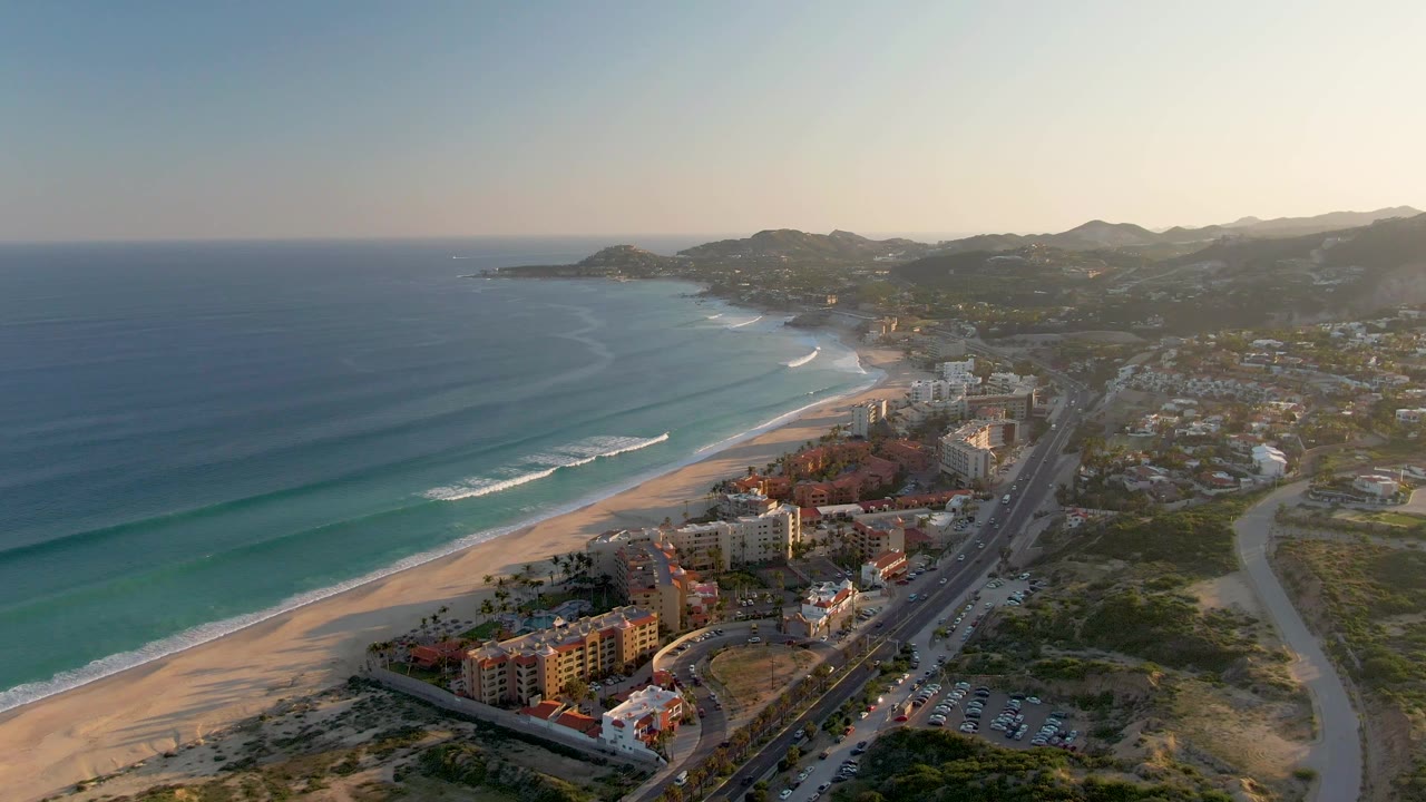 Aerial view over San Jose del Cabo coastline at 0:00, with blue ocean waves approaching a sandy beach, shoreline buildings, a road, distant hills, and no visible people.
