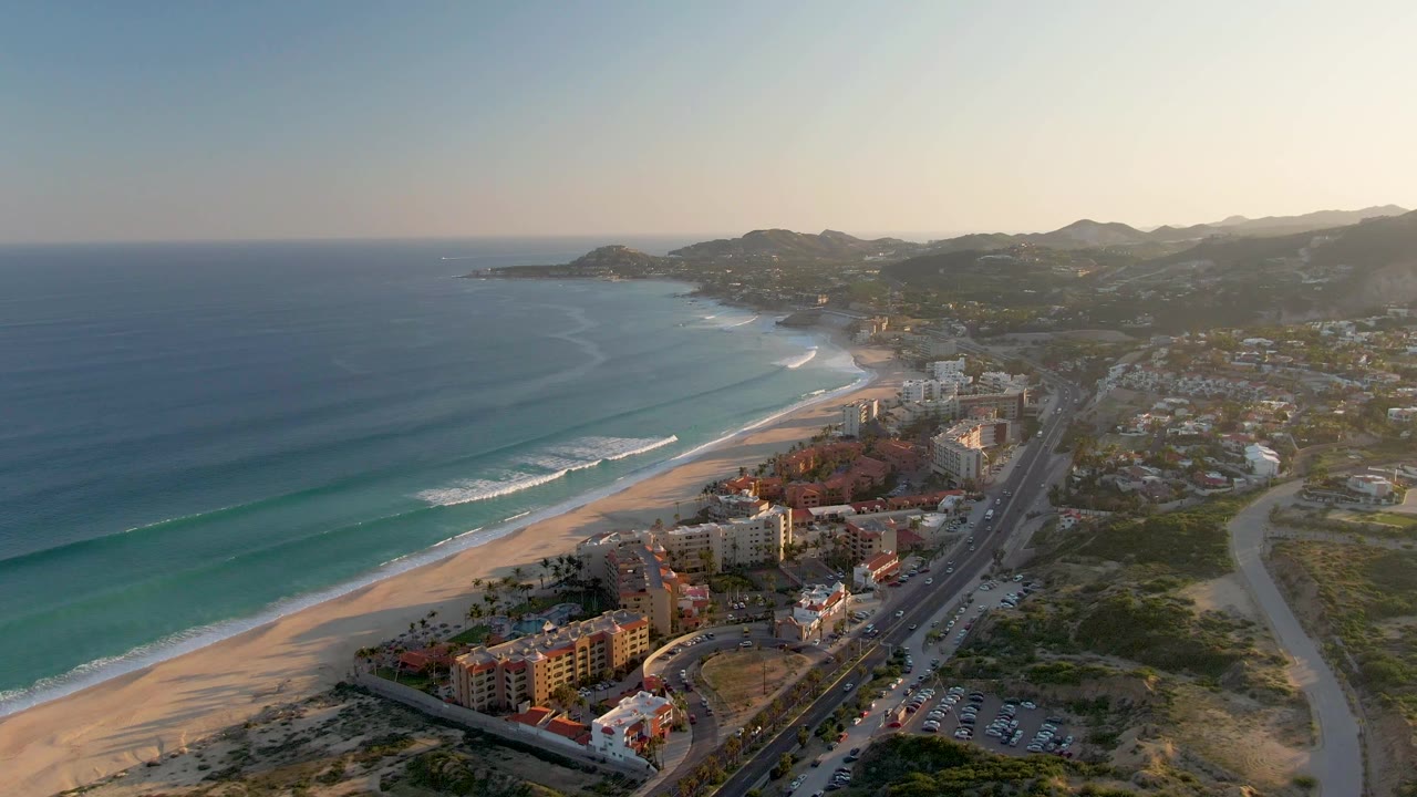Aerial frame from San Jose del Cabo at 0:02, with green-blue water, white wave breaks, a broad sand strip, beachfront buildings, a parallel road, low mountains, and no visible people.