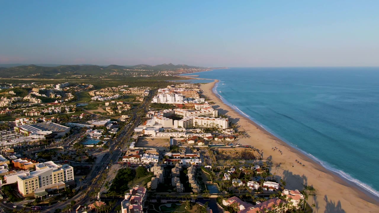 Aerial view over San Jose Del Cabo with low-rise buildings and a coastal road beside a sandy beach, blue water to the right, distant hills on the horizon, and no visible people.