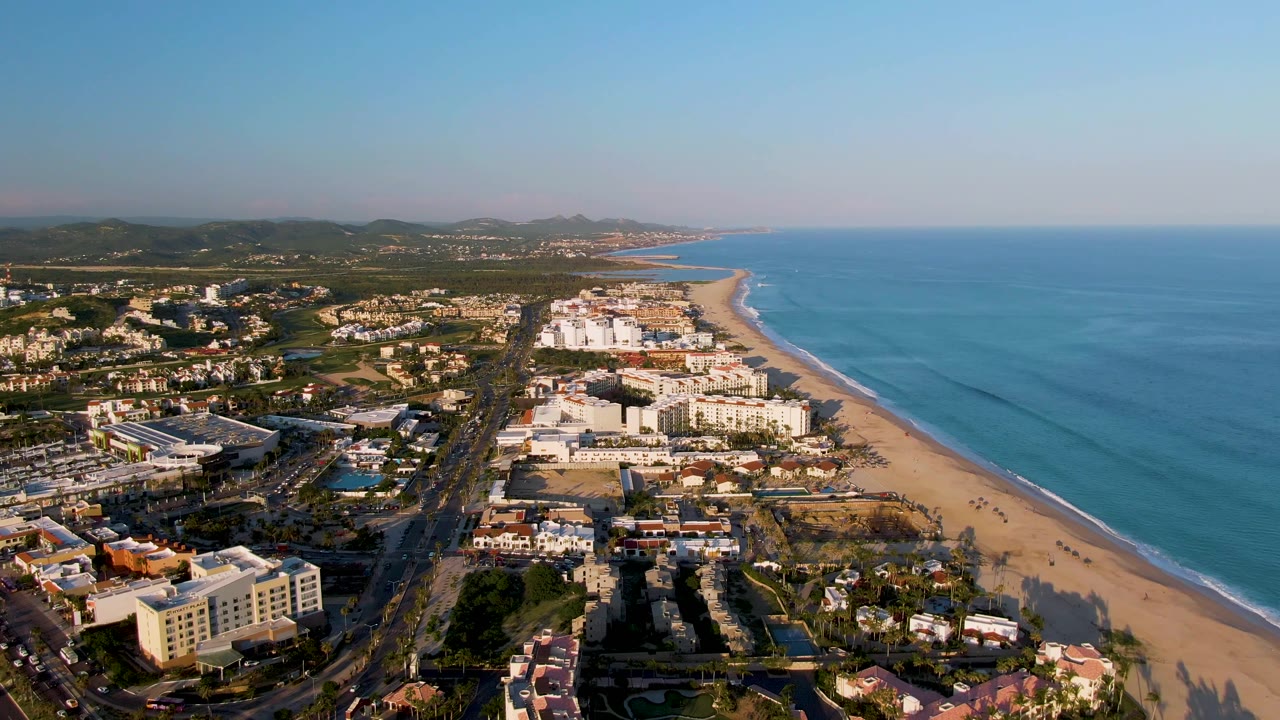 High oblique aerial over San Jose Del Cabo showing a developed beachfront district, a straight coastal road, sandy shoreline, calm blue water, inland hills, and no visible people.